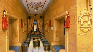 A corridor with ornate walls, a ceiling fan, and a row of blue cushioned stools. - Hotel Moonlight (Jaisalmer)