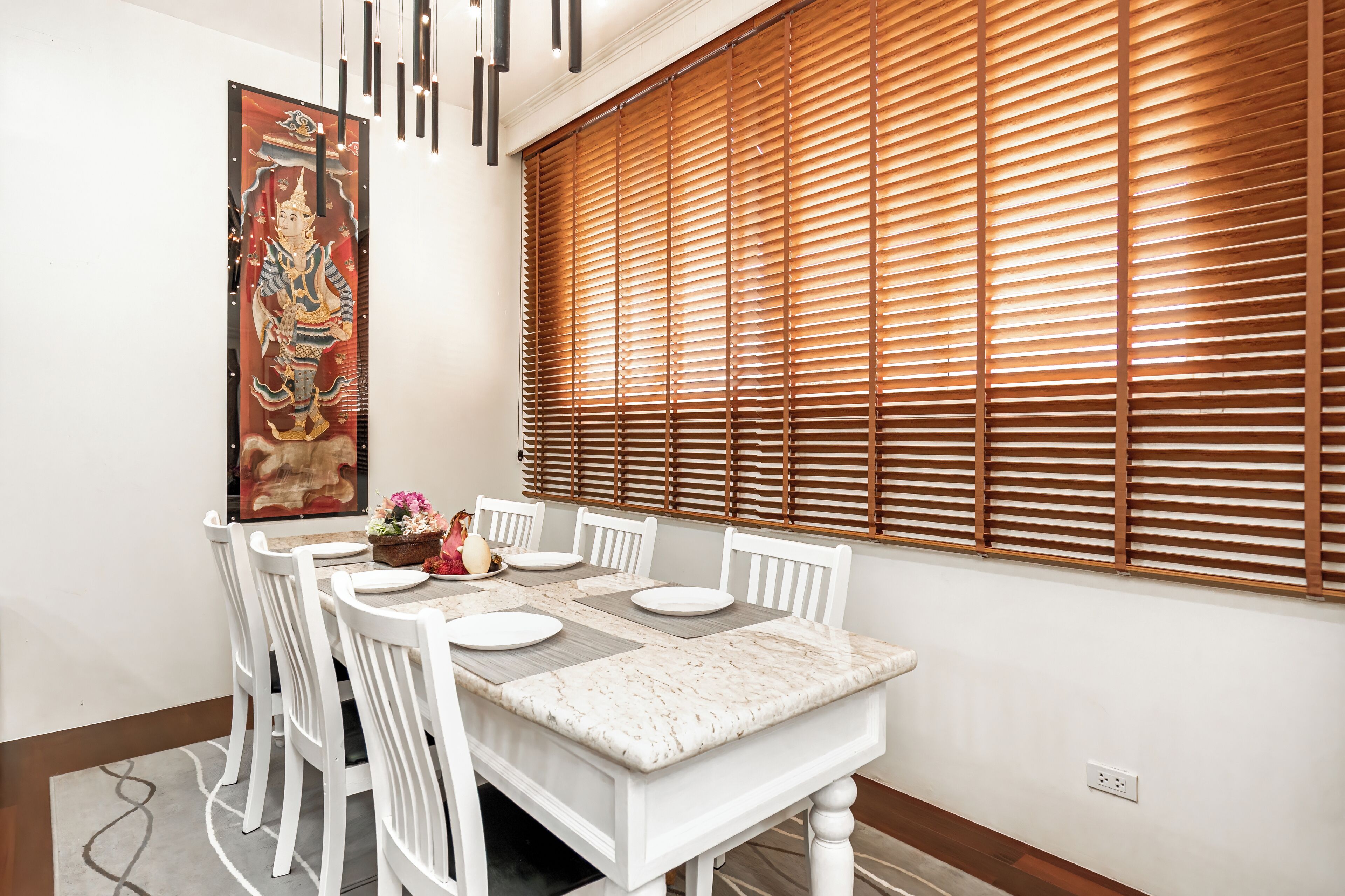 Un comedor con una mesa blanca dispuesta para seis comensales, sillas blancas, una persiana de madera y una obra de arte grande enmarcada en la pared.