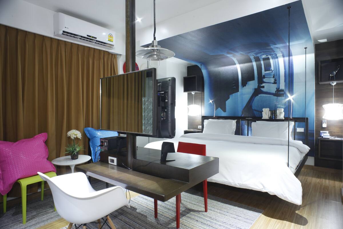 Quarto de hotel moderno com uma cama grande, uma escrivaninha com computador, uma cadeira, uma TV e um mural na parede.