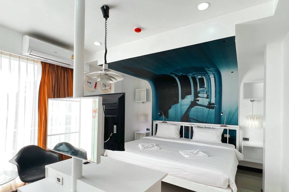 Quarto de hotel com cama, mesa de cabeceira, escrivaninha, cadeira e uma janela grande com cortinas.