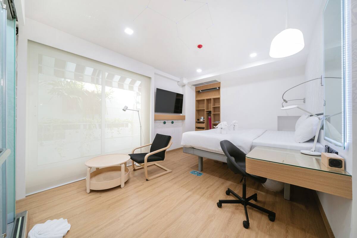 Quarto de hotel moderno com cama, escrivaninha, cadeira e box de vidro.