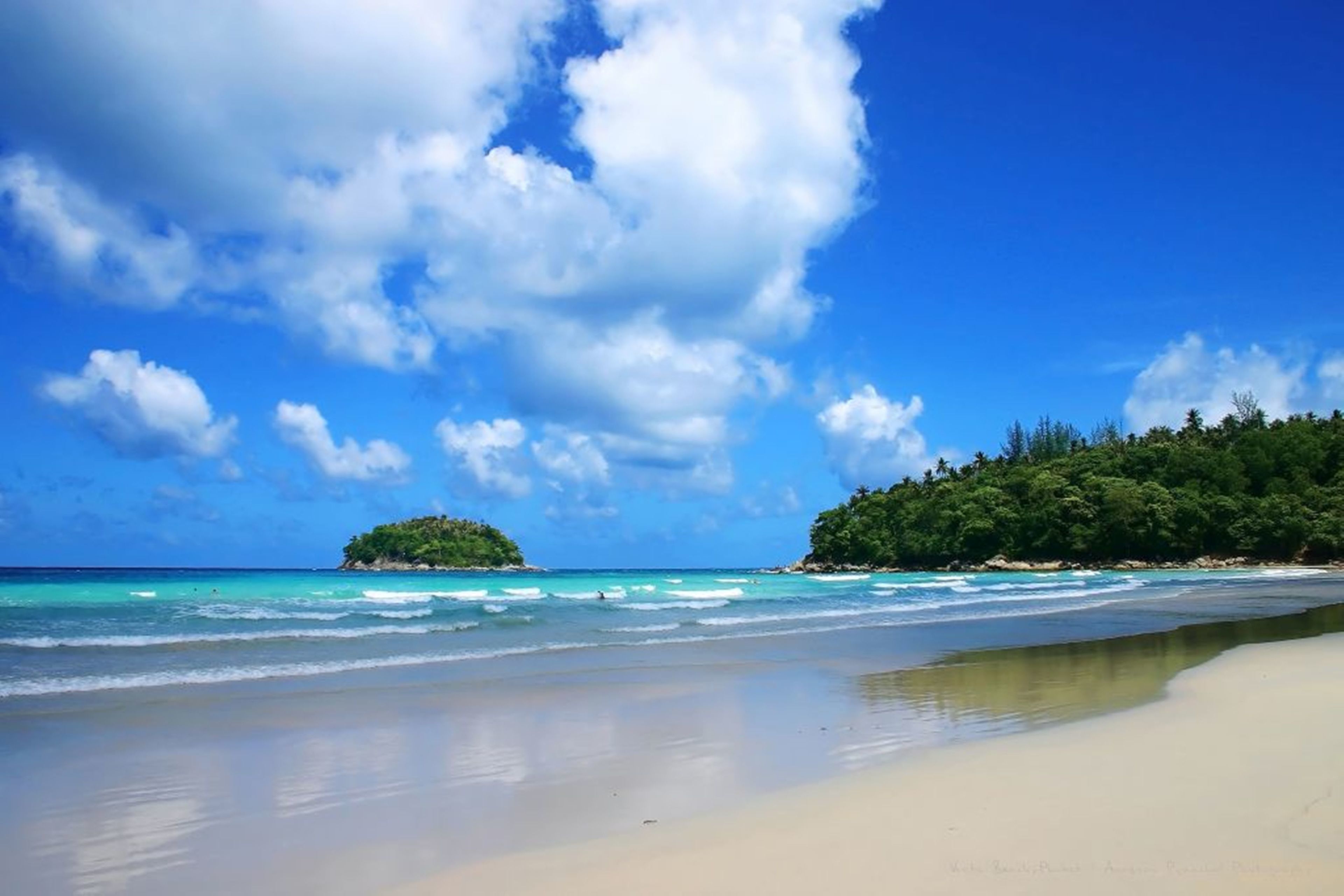 A beach with clear turquoise waters, a sandy shore, and a forested island in the distance.