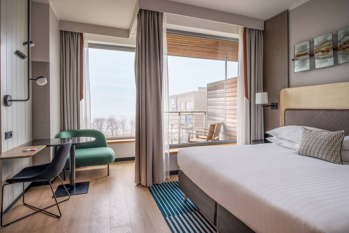Ein Hotelzimmer mit einem großen Bett, einem Schreibtisch mit Stuhl, einer grünen Couch und Blick ins Freie.
