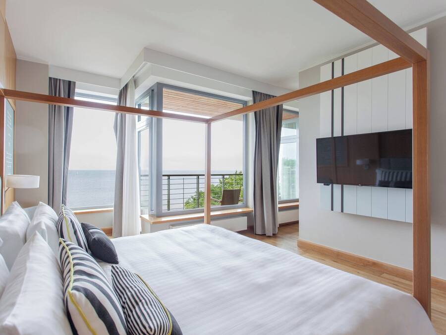 Quarto de hotel moderno com uma cama grande, uma TV fixada na parede e uma varanda com vista para o mar.