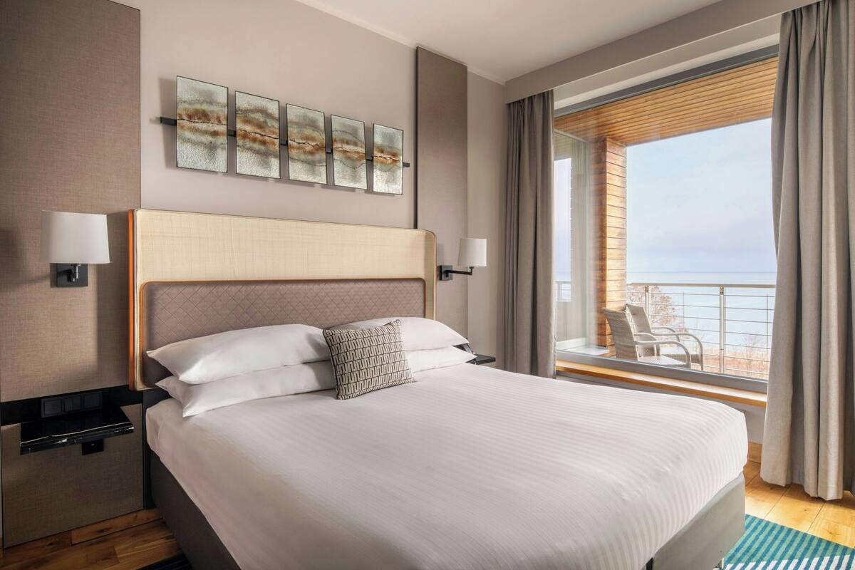 Ein Hotelzimmer mit einem großen Bett, Meerblick und einem Balkon mit Sitzgelegenheit.