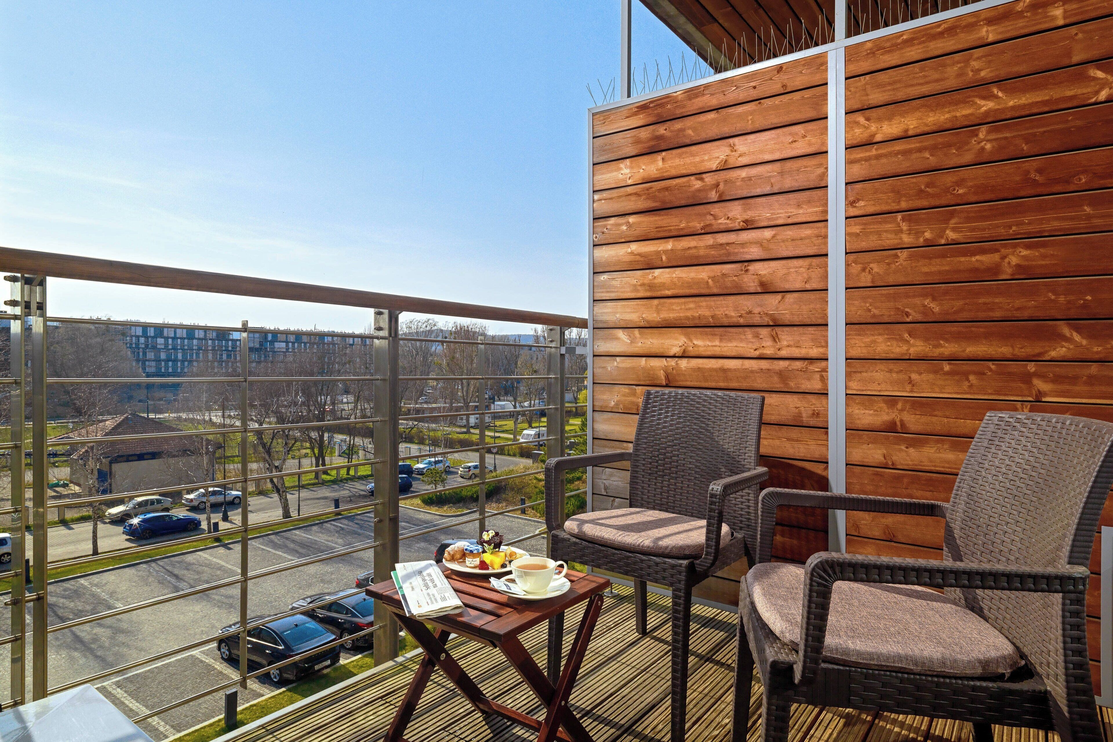 Ein Balkon mit Korbstühlen, einem Tisch mit einer Speiseplatte und einer Tasse, und Blick auf einen Parkplatz und Gebäude.