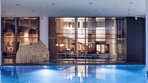 Un área de piscina interior moderna con un muro de vidrio transparente, una formación rocosa y un espacio de estar con mesas y sillas.