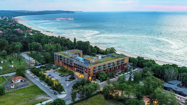 Un edificio moderno con techo verde, ubicado cerca de una playa con muelle y una extensa masa de agua.
