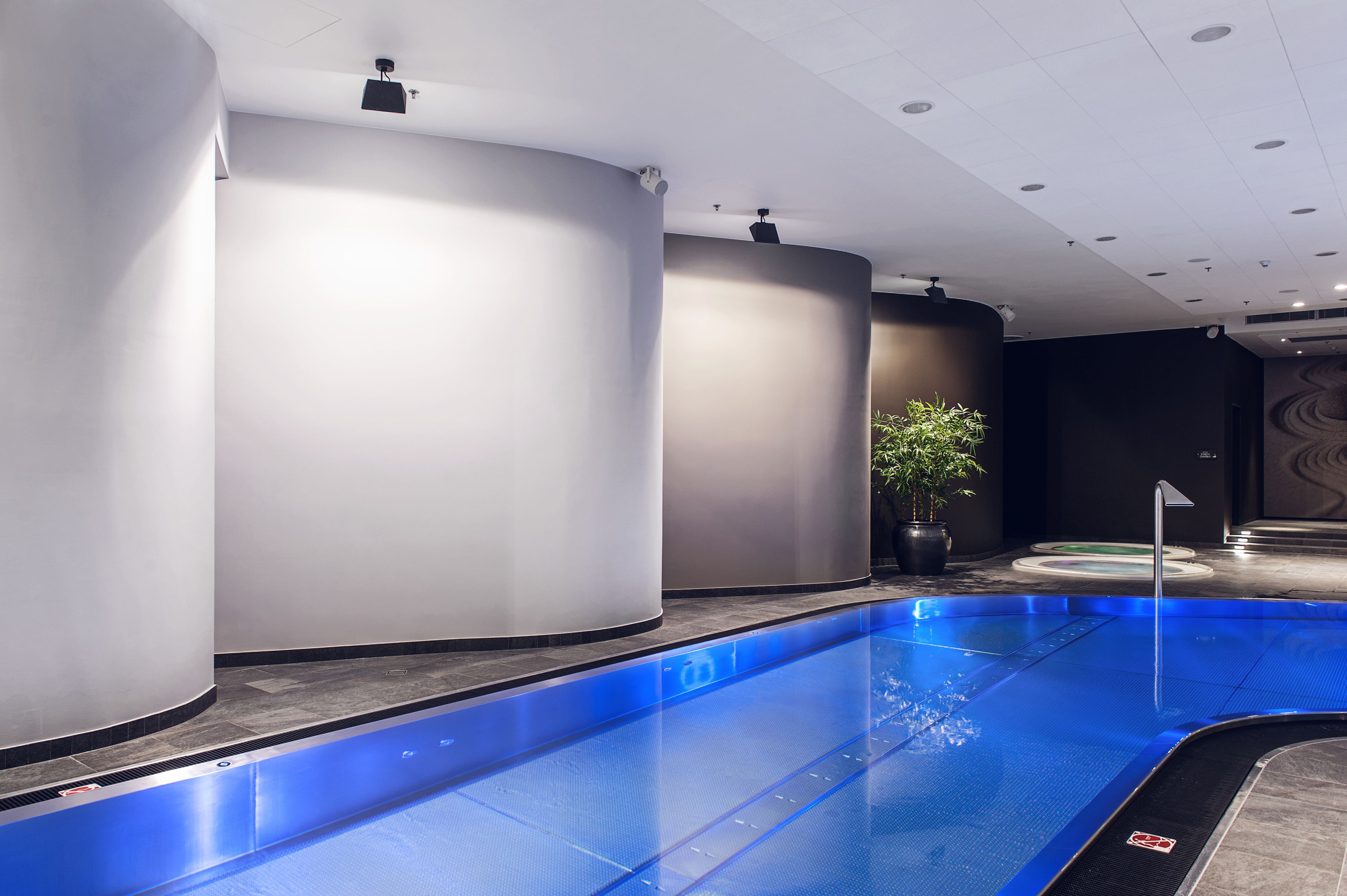 Una piscina interior moderna con un borde iluminado en azul, grandes columnas cilíndricas y una planta en maceta.