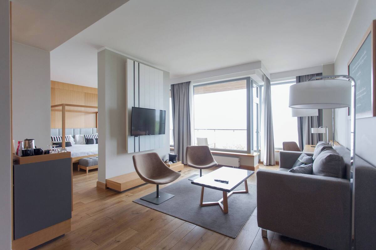Ein modernes Hotelzimmer mit einem Bett, zwei Sesseln, einem Sofa, einem Fernseher und einem großen Fenster.