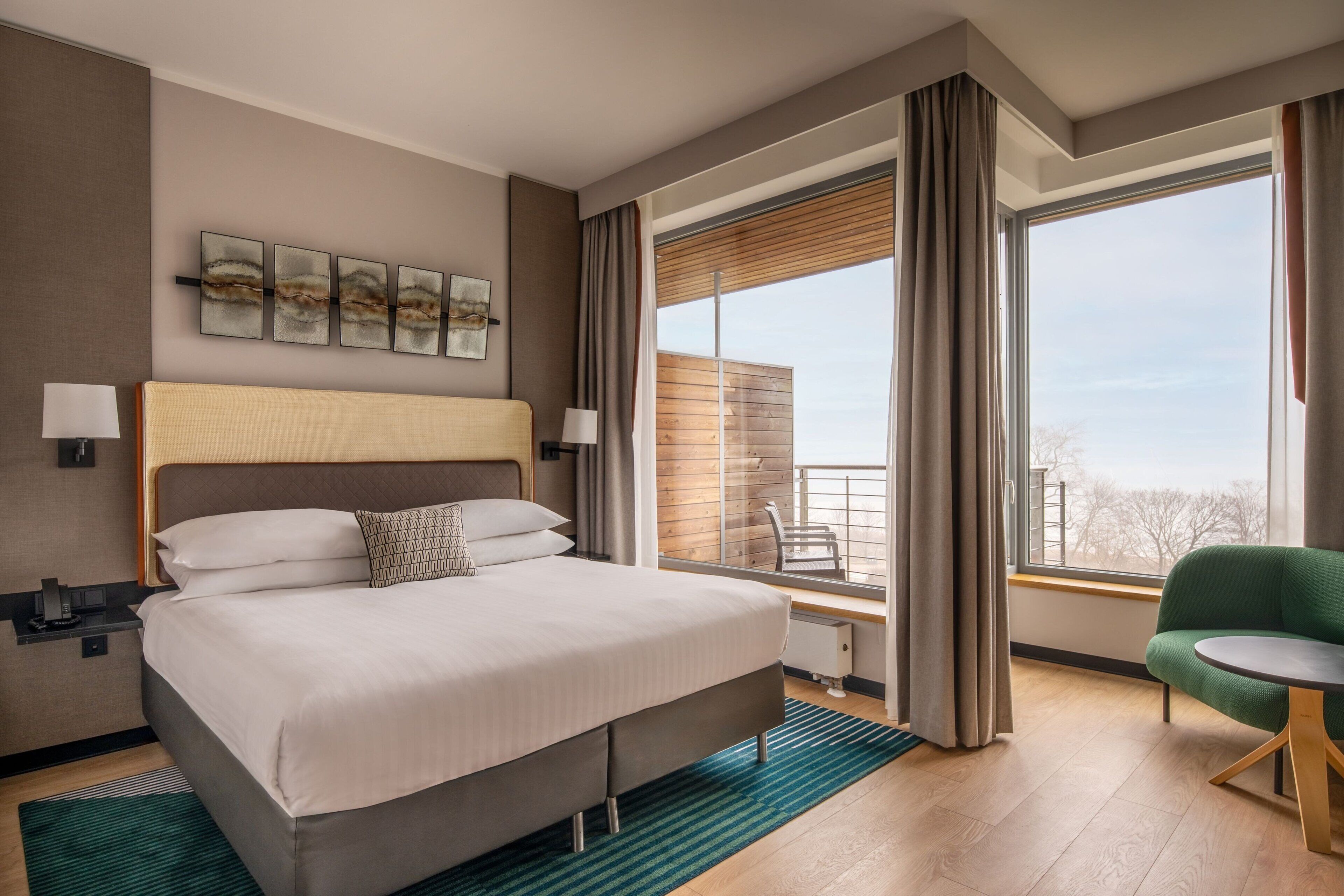 Quarto de hotel moderno com uma cama grande, uma poltrona verde e vista da cidade através de amplas janelas.