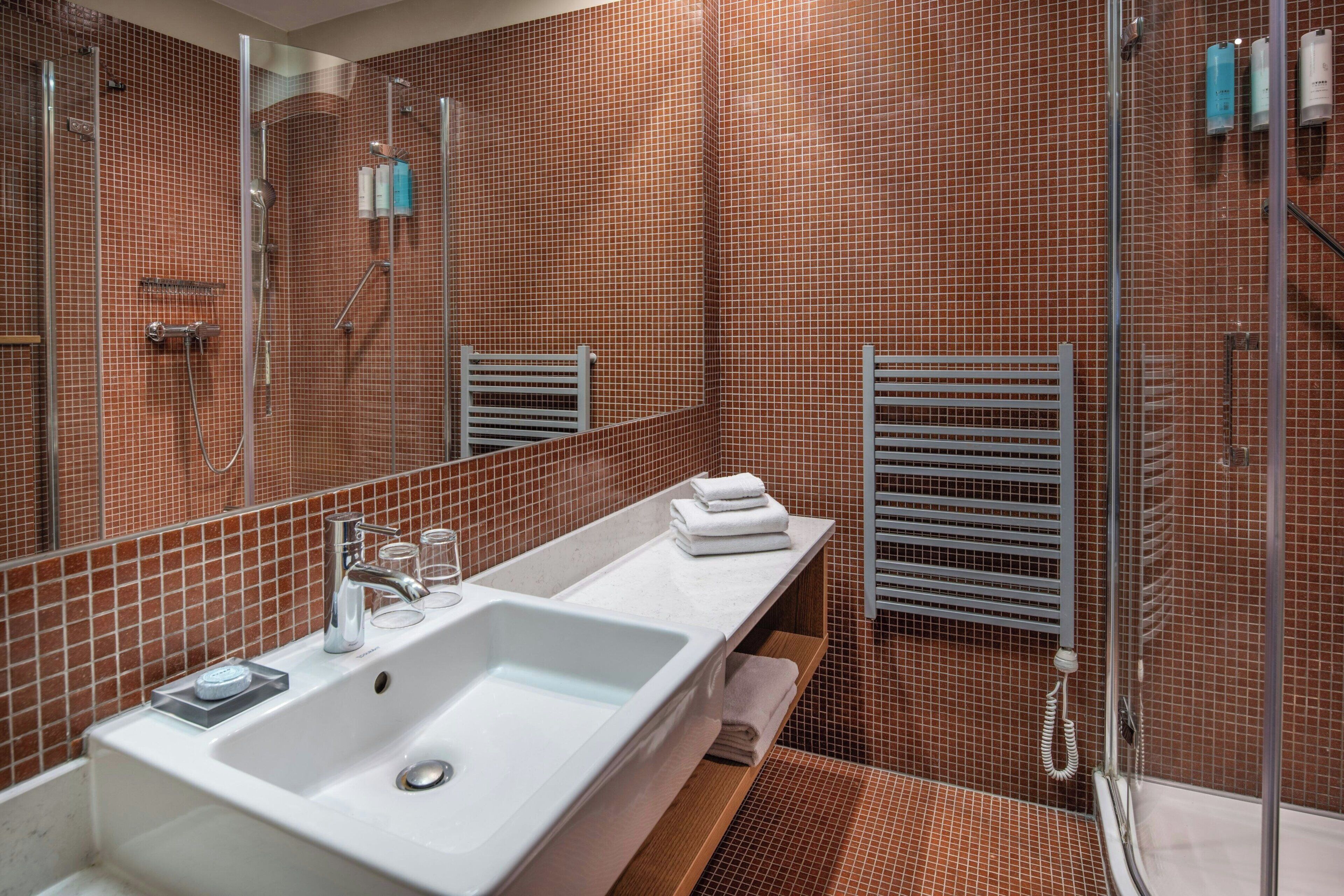 Un baño moderno con un lavamanos grande de color blanco, ducha con mampara de vidrio y pared con azulejos.