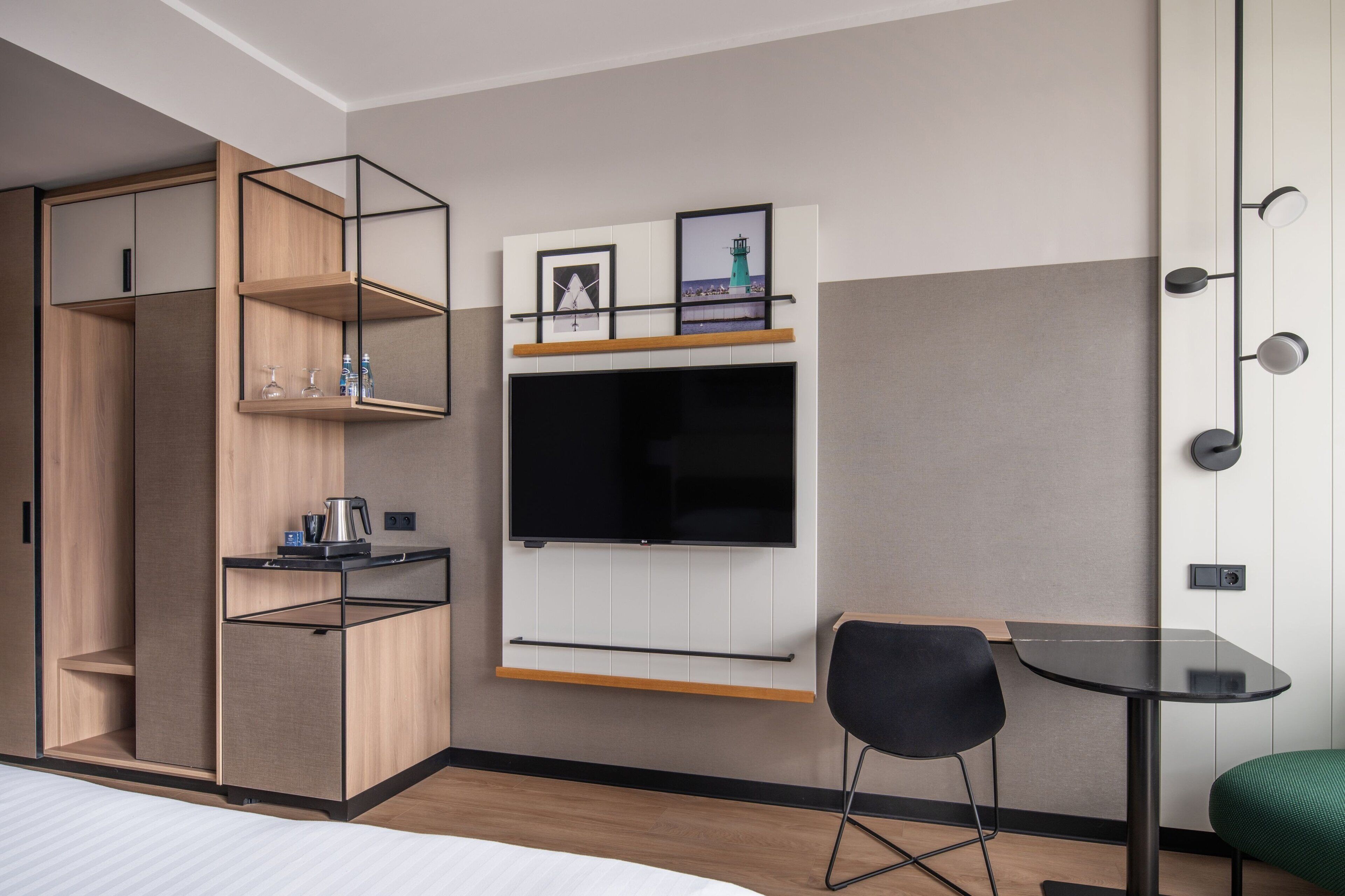 A modern hotel room with a bed, a TV mounted on the wall, a small table, a chair, and a green ottoman.