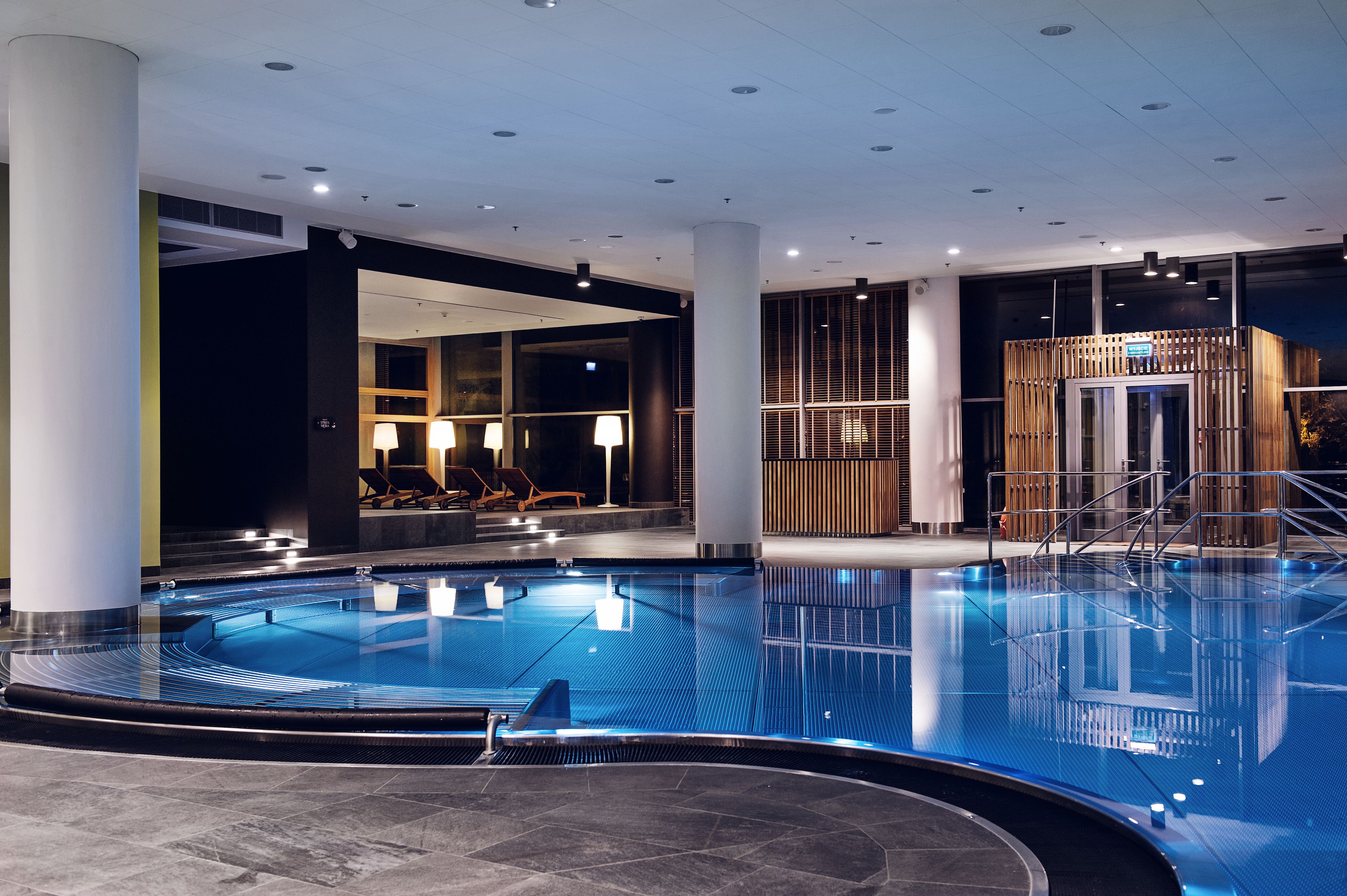 A modern indoor swimming pool with clear water, surrounded by a spacious area with seating and a reception desk.