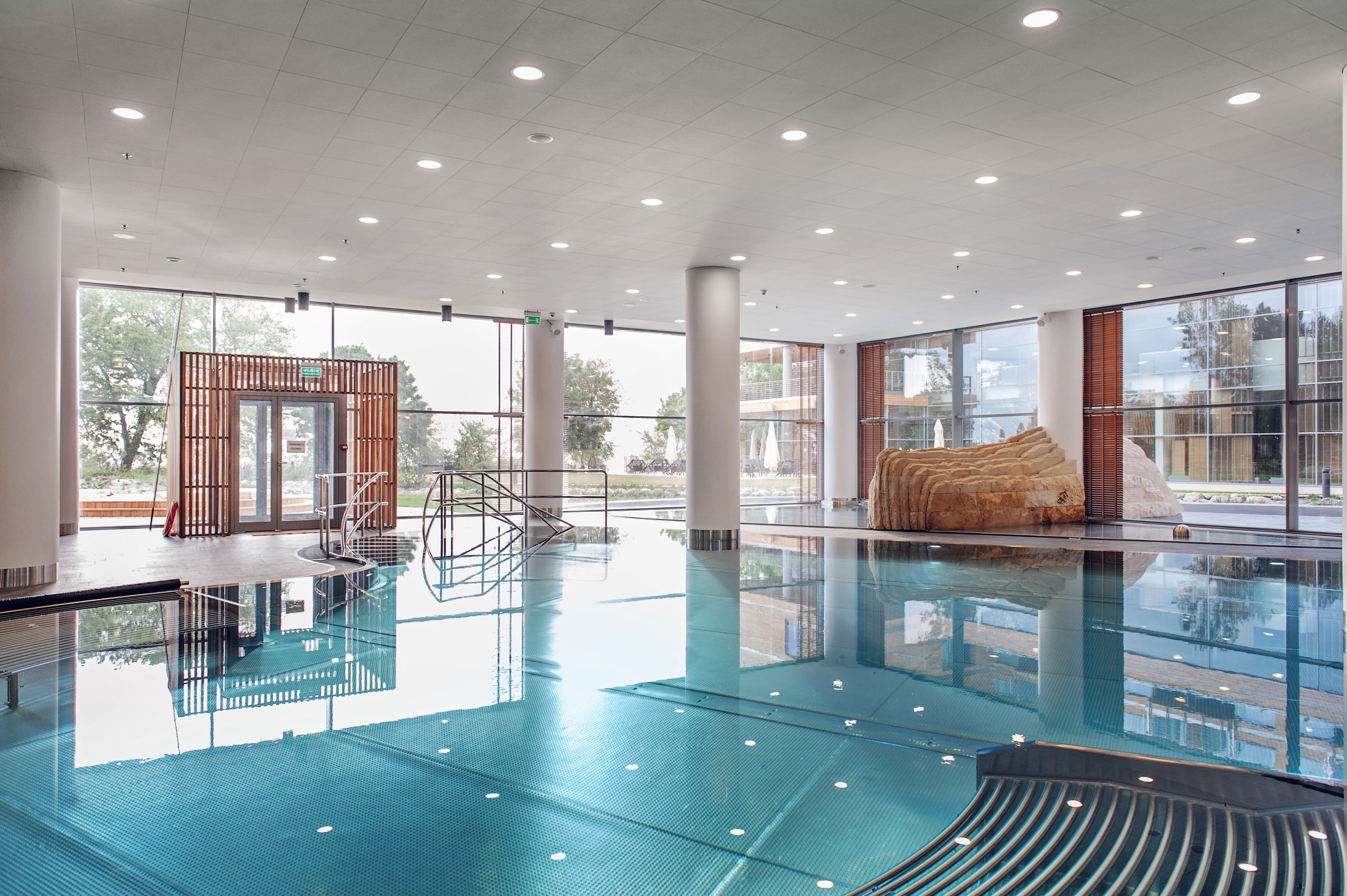 A spacious indoor swimming pool with clear glass walls, a wooden structure, and a large rock feature.