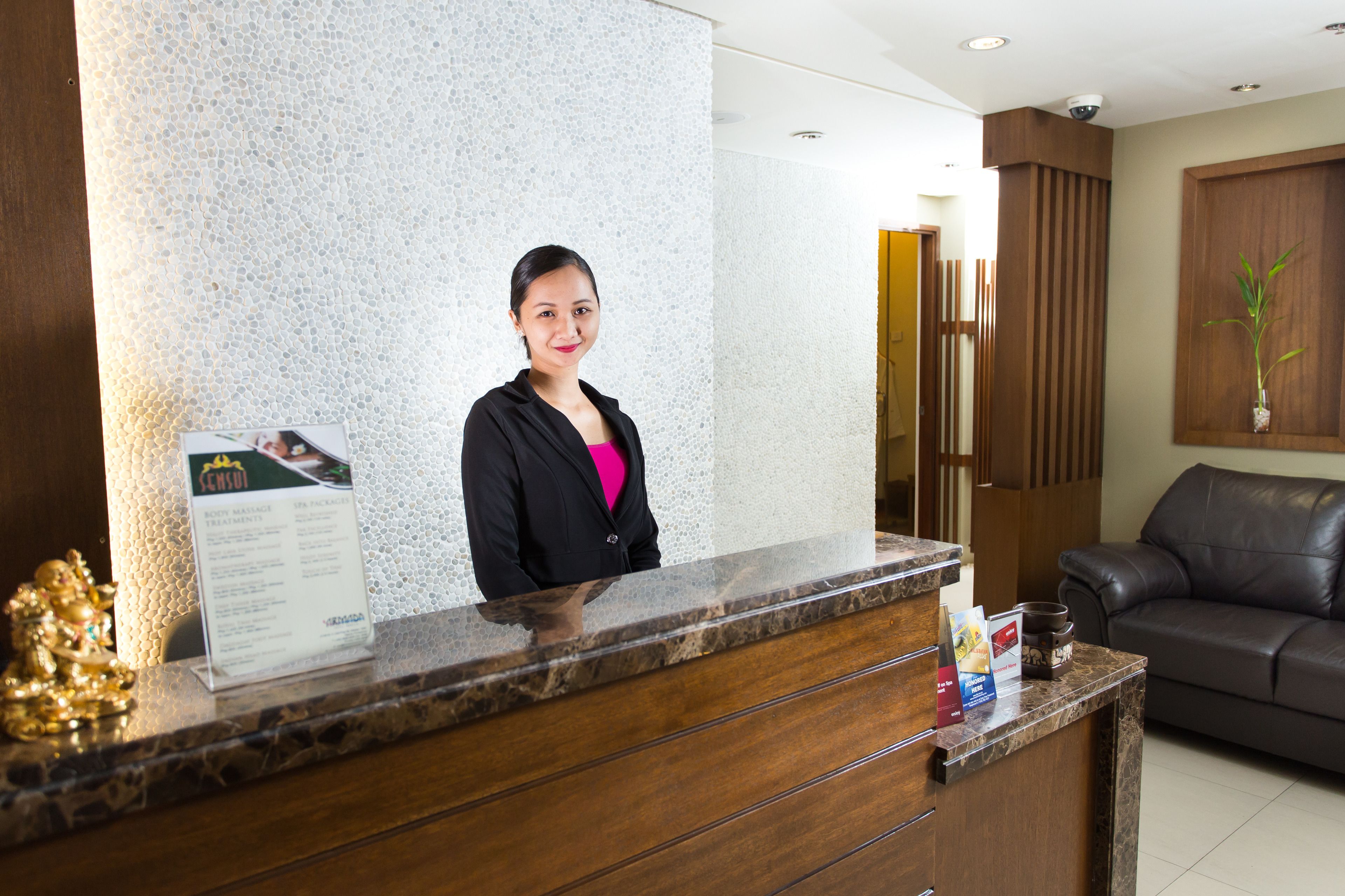 A reception desk with a woman standing behind it, a signboard, a brown leather sofa, and a potted plant.