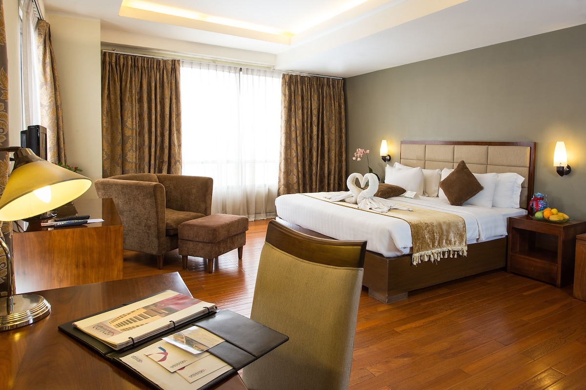 Quarto de hotel com cama, escrivaninha com luminária, cadeira, mesa de cabeceira e apoio para frutas.