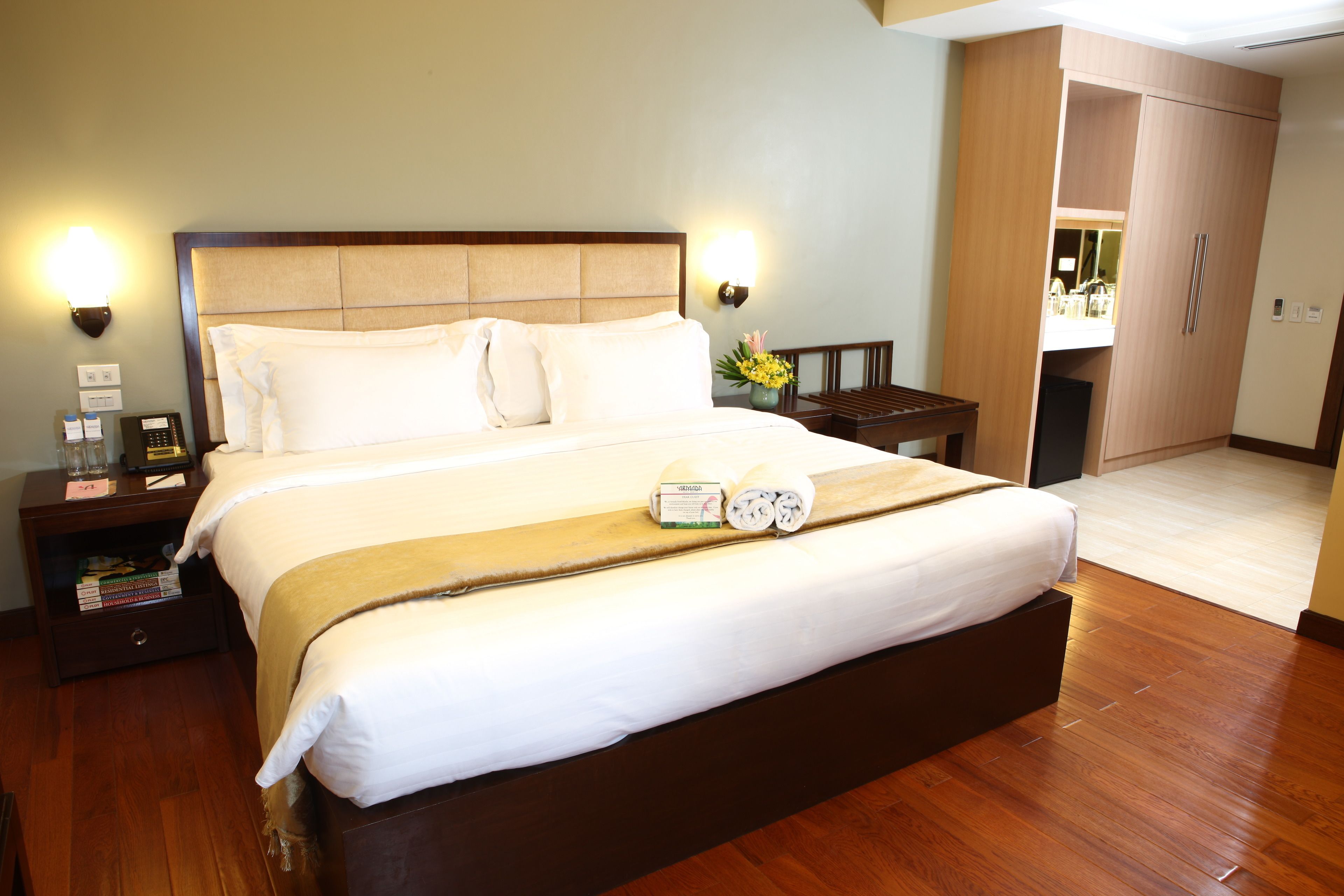 A hotel room with a large bed, wooden headboard, bedside tables with lamps, a nightstand with a telephone, and a view of the bathroom and kitchen through an open door.