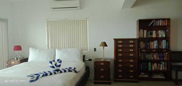 A hotel room with a bed, a bedside lamp, a bookshelf, and a window with blinds.