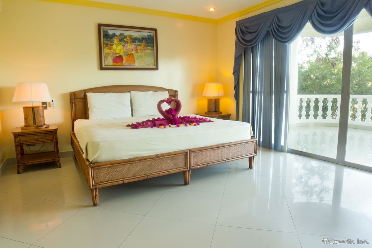 Habitación de hotel con una cama grande, dos mesitas de noche con lámparas, un cuadro en la pared y un balcón con barandilla.