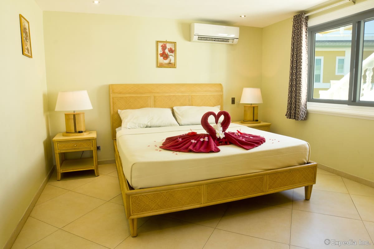 Una cama bien hecha con una toalla con forma de corazón y flores, dos lámparas de noche, un cuadro en la pared y un equipo de aire acondicionado.