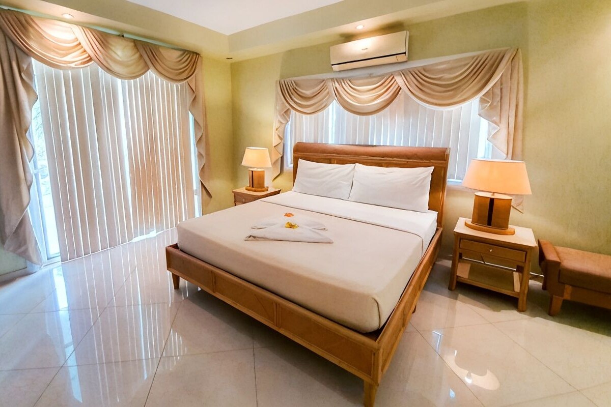 A hotel room with a large bed, two bedside tables with lamps, a bench, and a window with curtains.