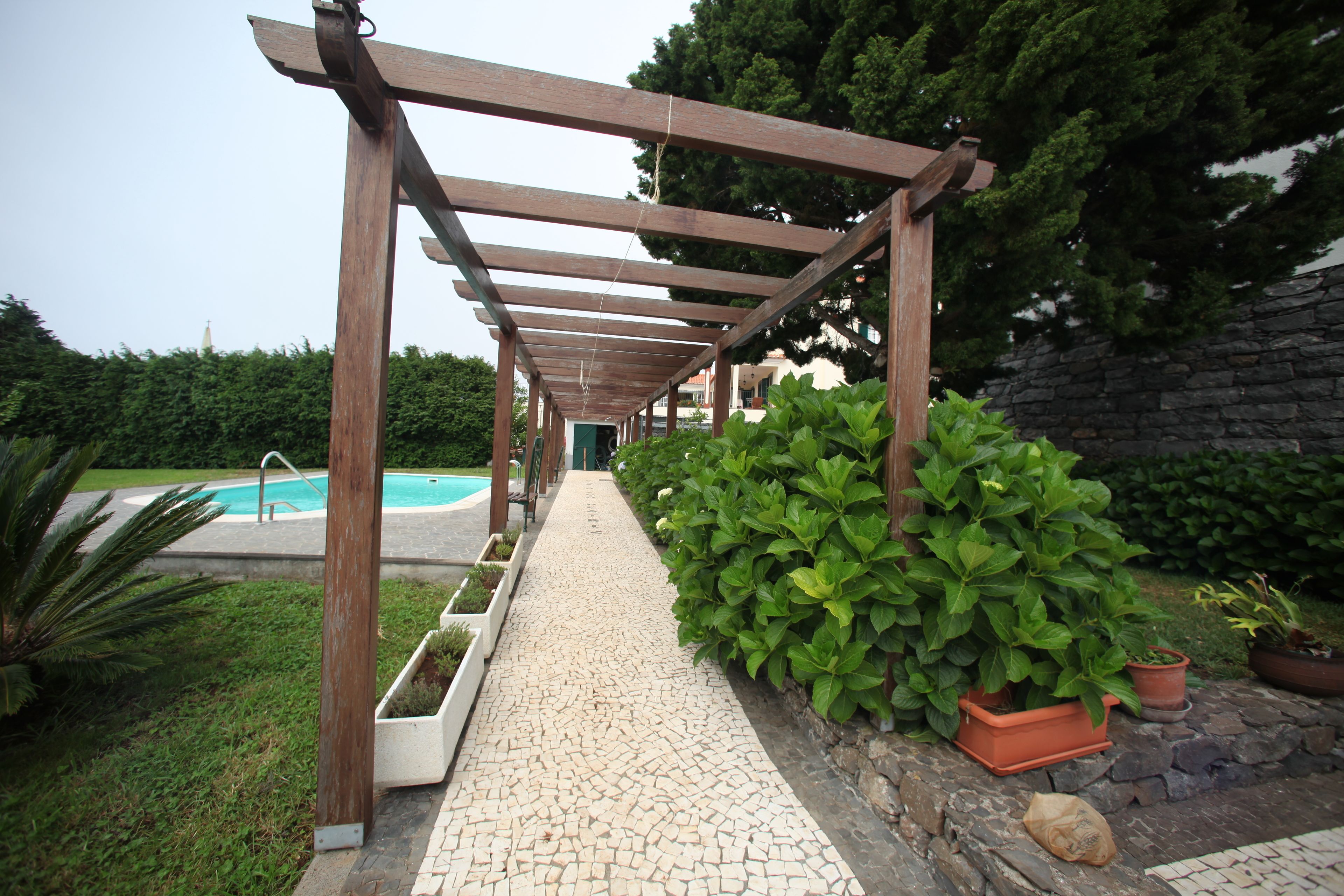 A covered walkway with a tiled floor, leading to a pool area, surrounded by greenery and stone walls.
