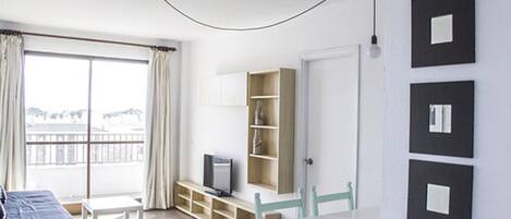 Quarto com uma grande janela, televisão sobre um suporte de madeira, uma mesa branca e uma cadeira. Há quatro quadros na parede.