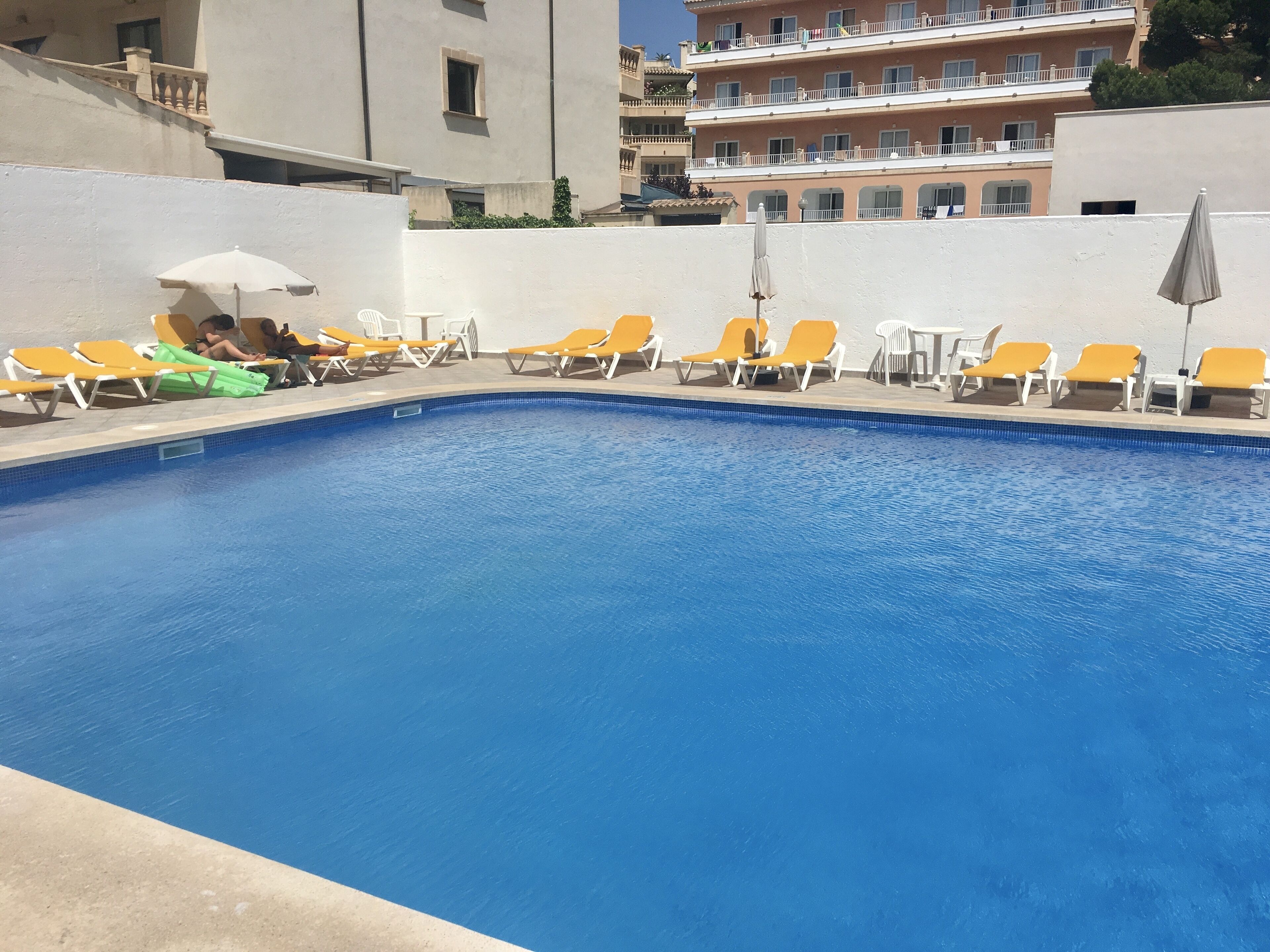 A swimming pool with yellow lounge chairs and umbrellas, surrounded by a white wall and buildings in the background.