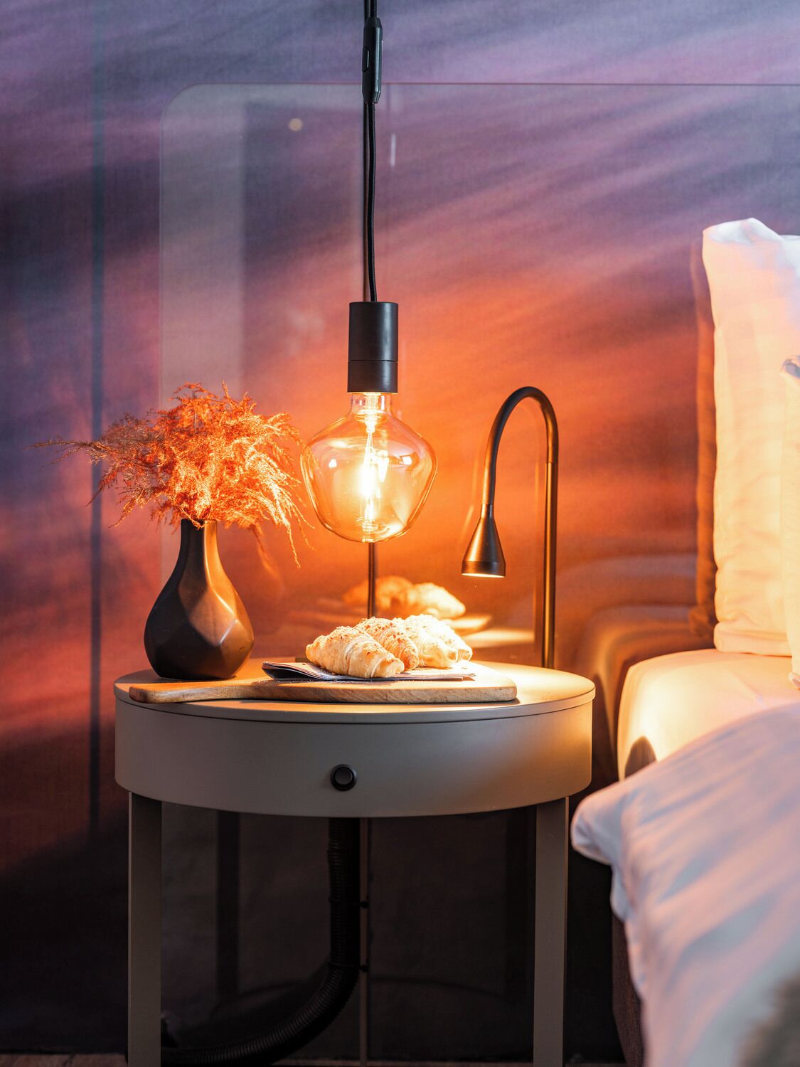 A bedside table with a lamp, a vase with dried flowers, and a plate with croissants.