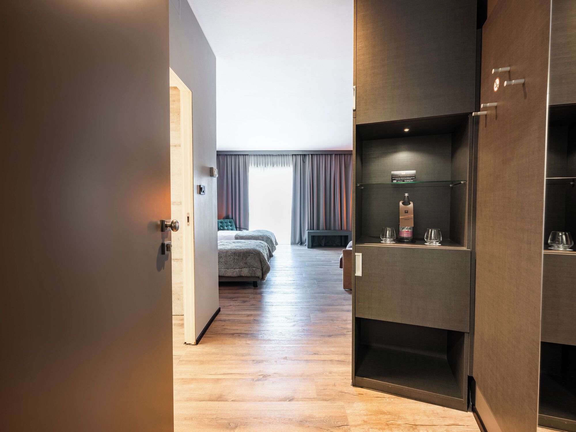 A hotel room with a wooden floor, a bed with a grey headboard, a bedside table, and a minibar with glasses and a bottle.