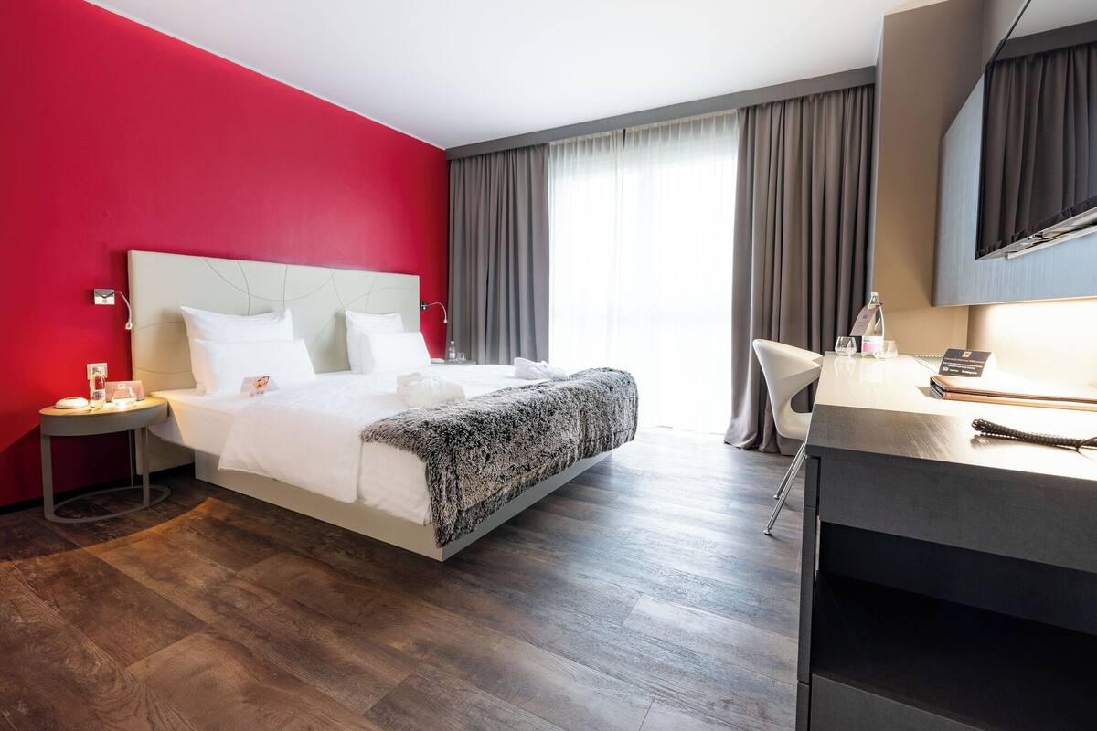 A modern hotel room with a red accent wall, a large bed with white linens, a desk with a chair, and a large window with curtains.