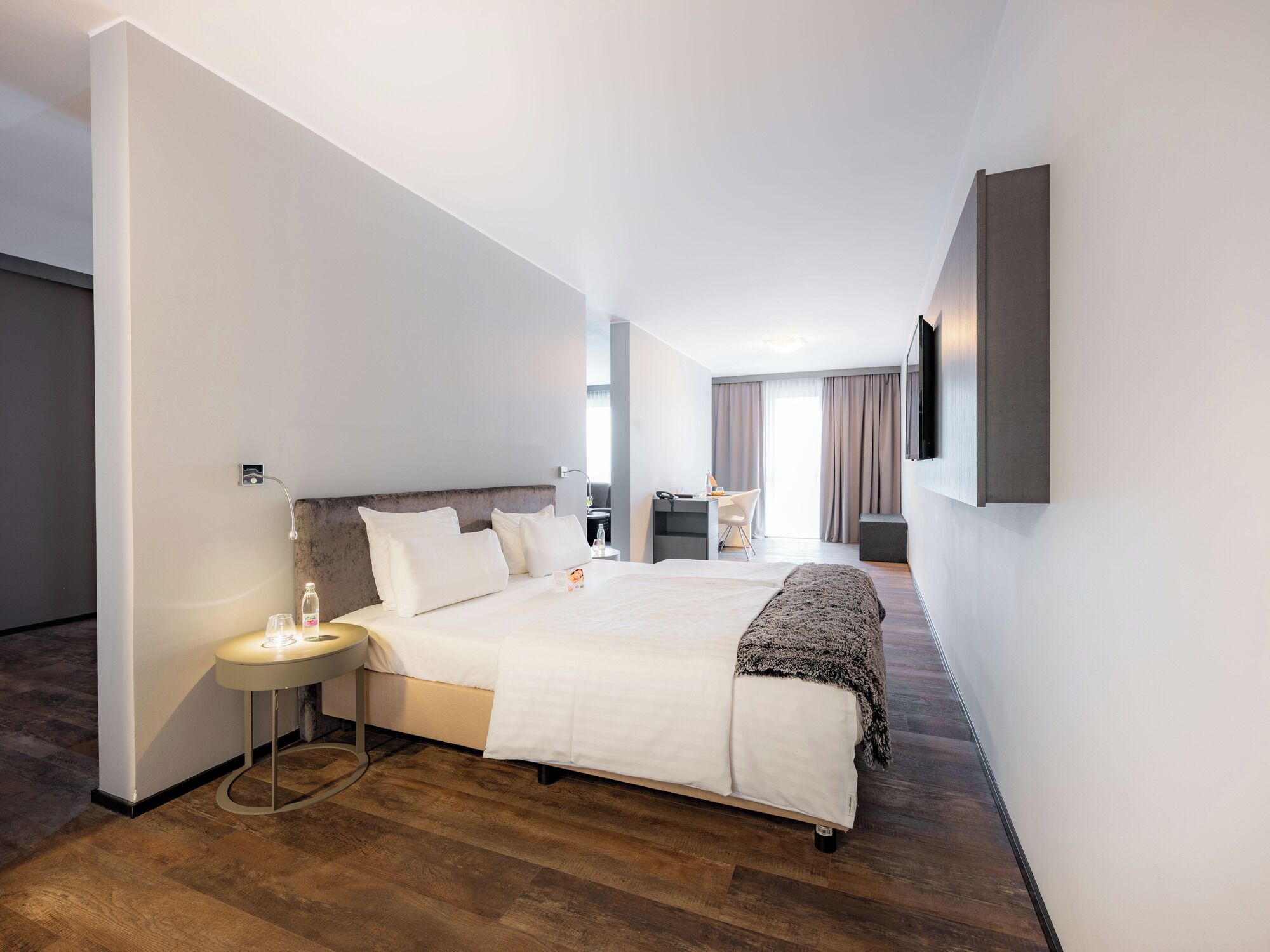 Une chambre d’hôtel moderne, dotée d’un grand lit, d’un bureau, d’une chaise, d’un téléviseur fixé au mur et d’une fenêtre avec des rideaux.