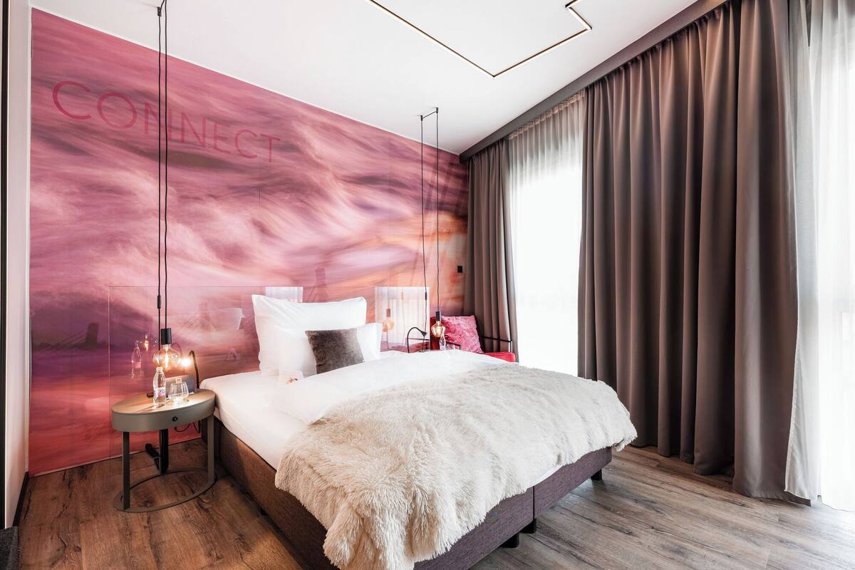 A modern bedroom with a large bed, a nightstand with a lamp, a wall mural, and a view of the outdoors.