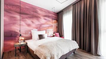 A modern bedroom with a large bed, a nightstand with a lamp, a wall mural, and a view of the outdoors.
