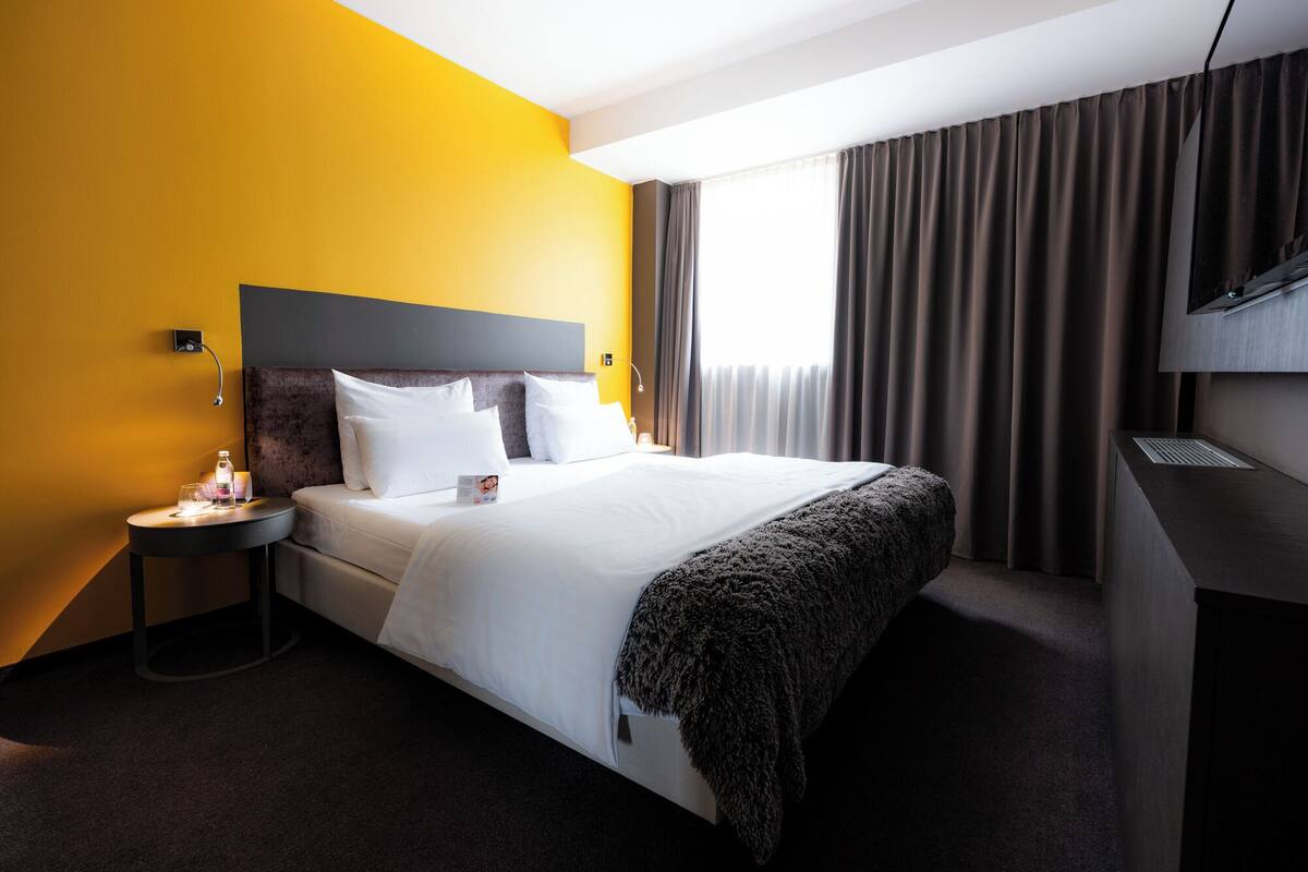 A hotel room with a bed, a bedside table, a lamp, a TV, and a window with curtains.