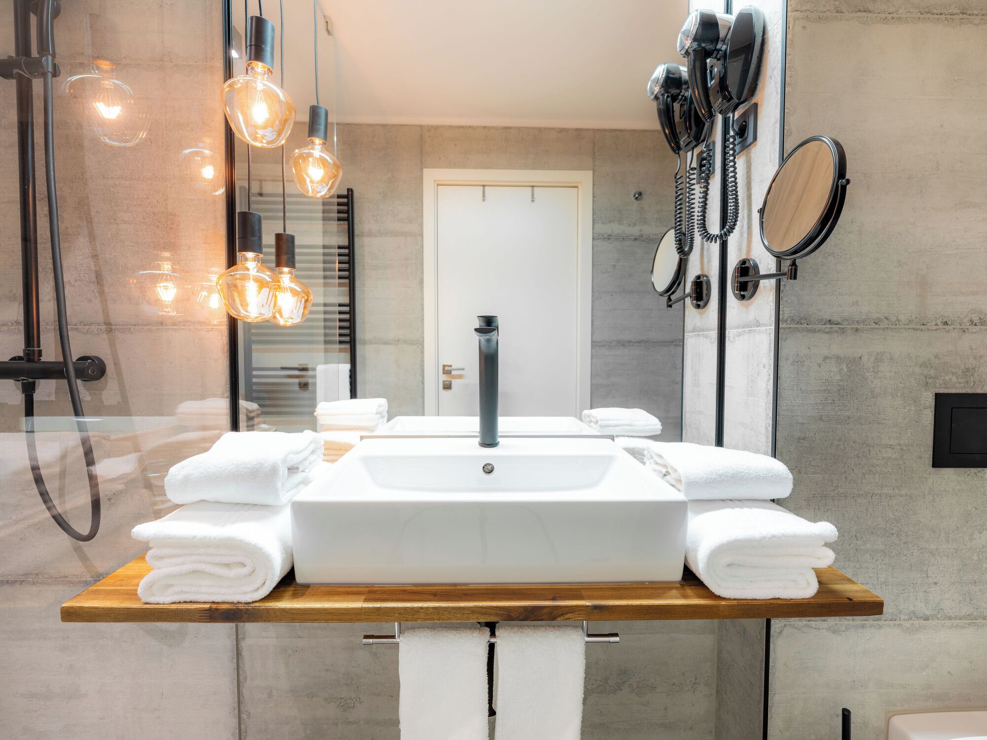 A modern bathroom with a large sink, multiple towels, a handheld showerhead, and a mirror with a round frame.