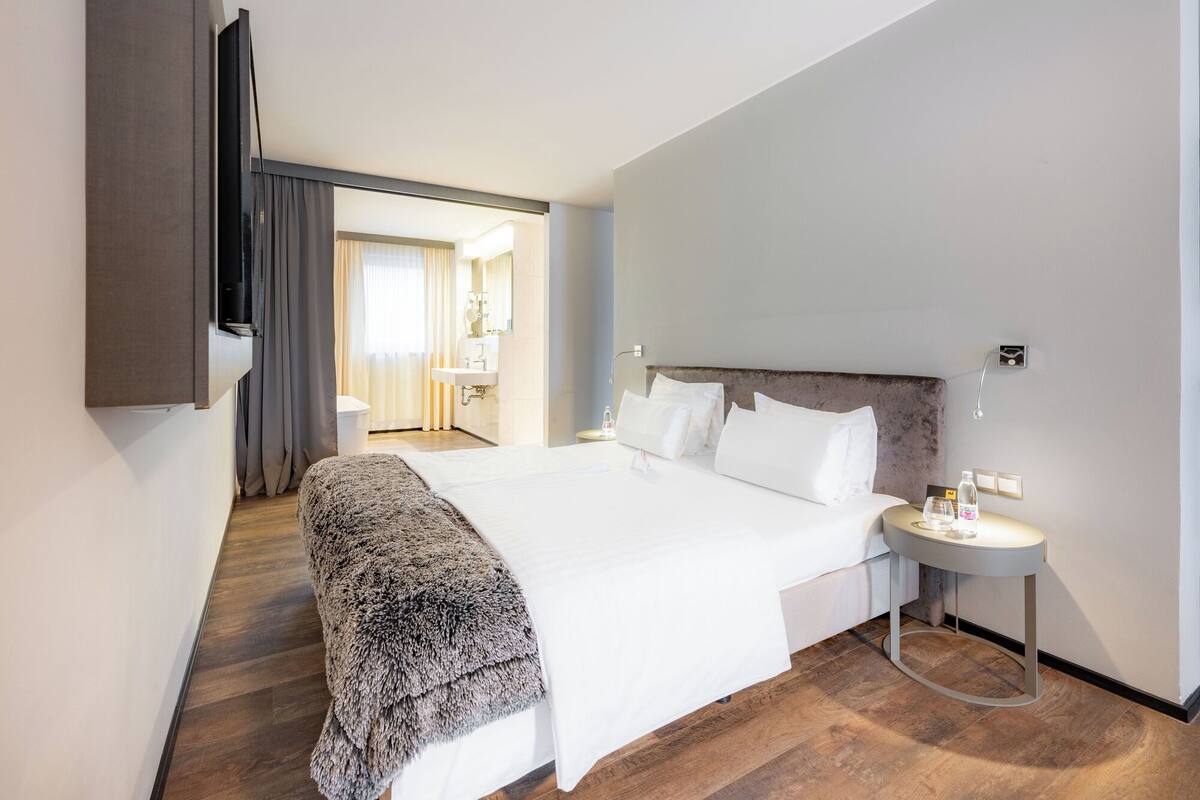A modern hotel room with a large bed, a bedside table with a lamp, and a view of the bathroom and hallway.
