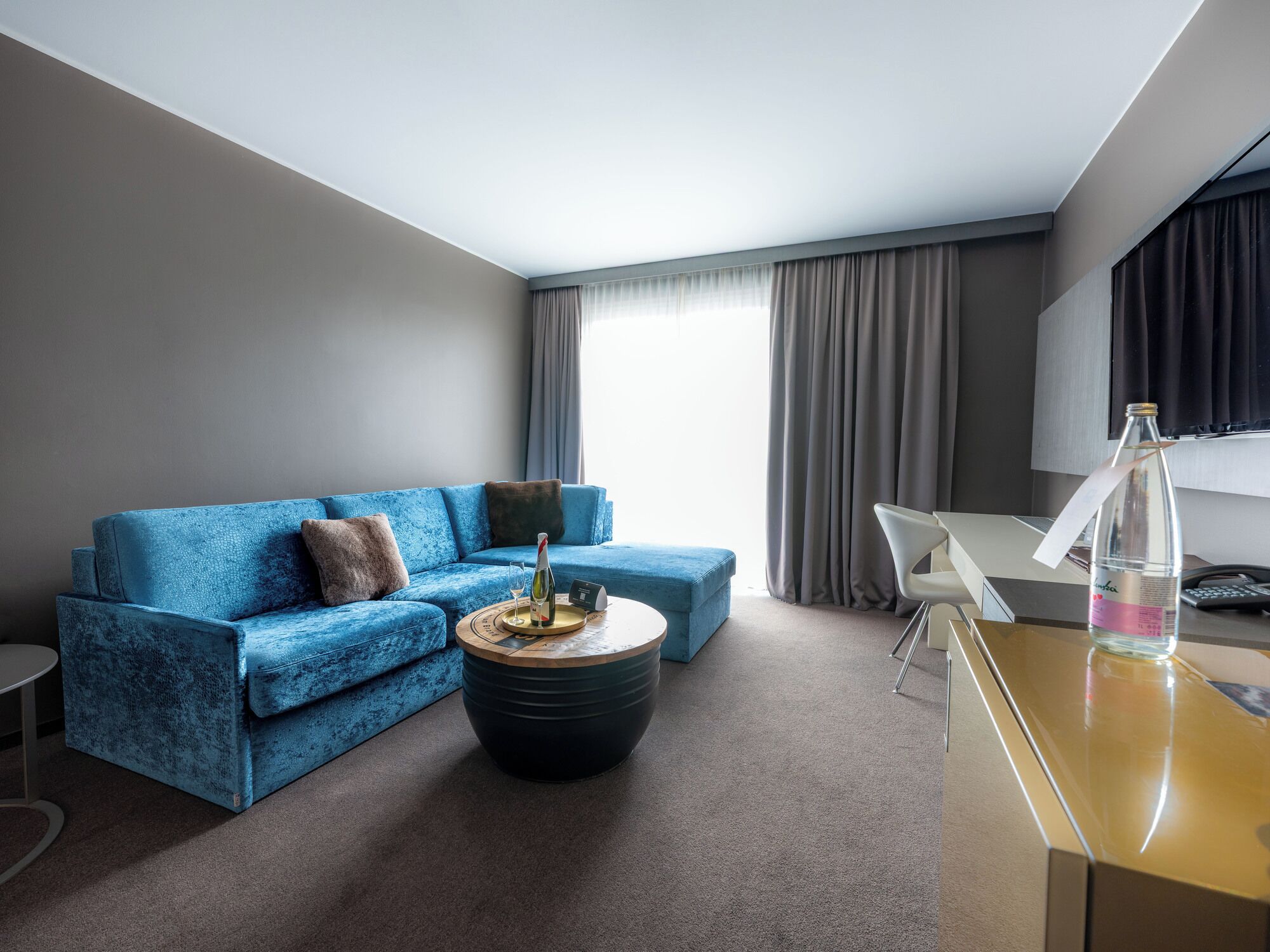 A modern hotel room with a blue sofa, a round coffee table, a desk with a chair, and a large window with curtains.