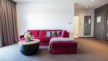 Uma sala de estar moderna com um sofá vermelho, uma mesa de centro redonda e um banquinho cinzento. Há uma porta à direita e uma janela com cortinas à esquerda.