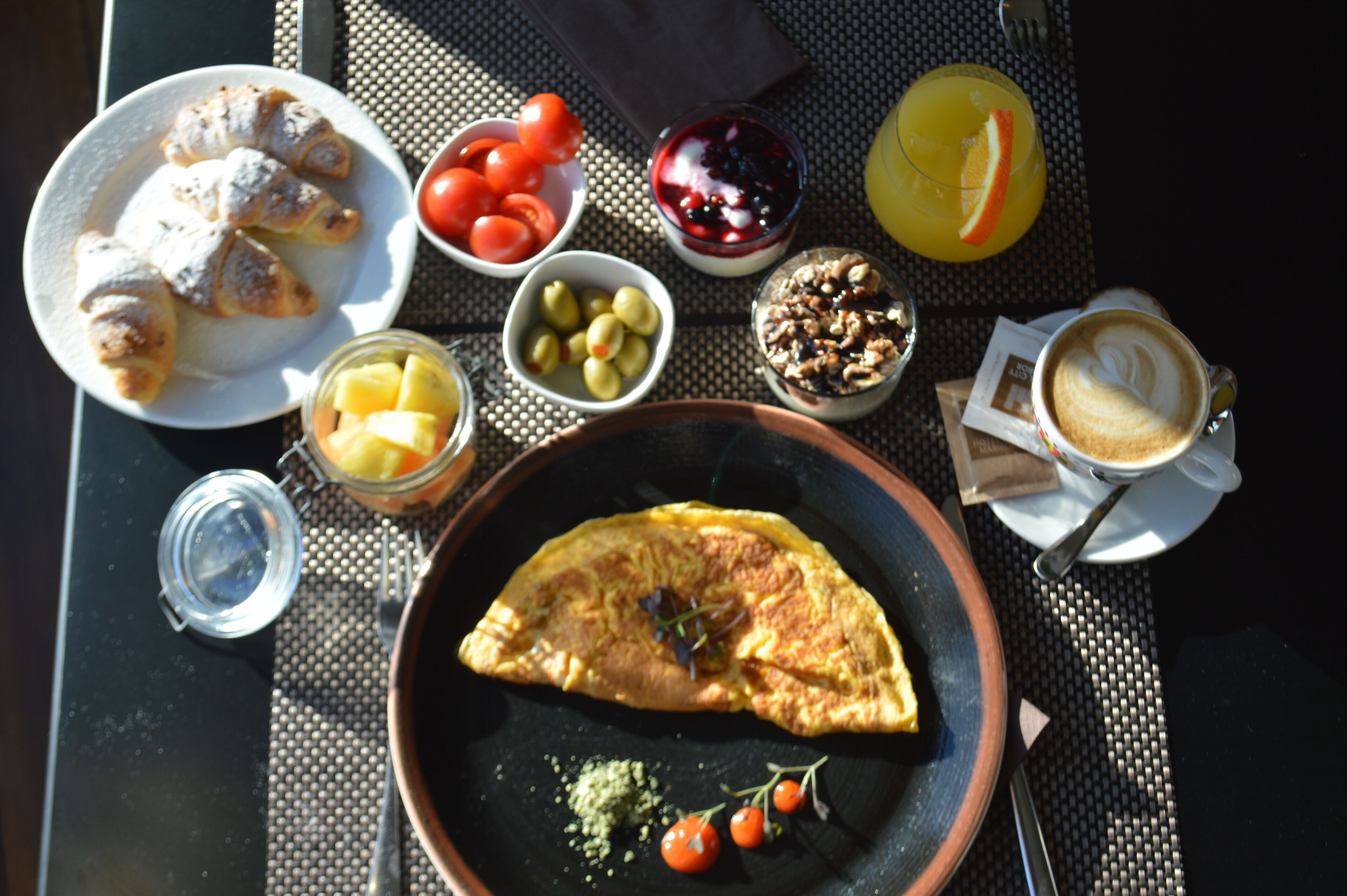 A table with a variety of breakfast items including croissants, tomatoes, olives, a fruit bowl, a bowl of yogurt with berries, granola, a glass of orange juice, and a cup of coffee.