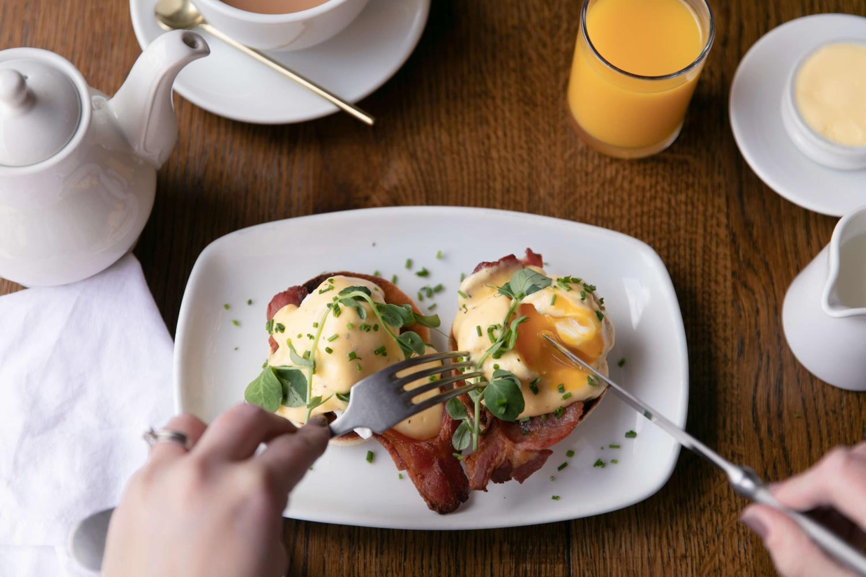 Uma mesa de café da manhã com um prato de ovos Benedict, um bule de chá, um copo de suco de laranja e uma xícara de café.