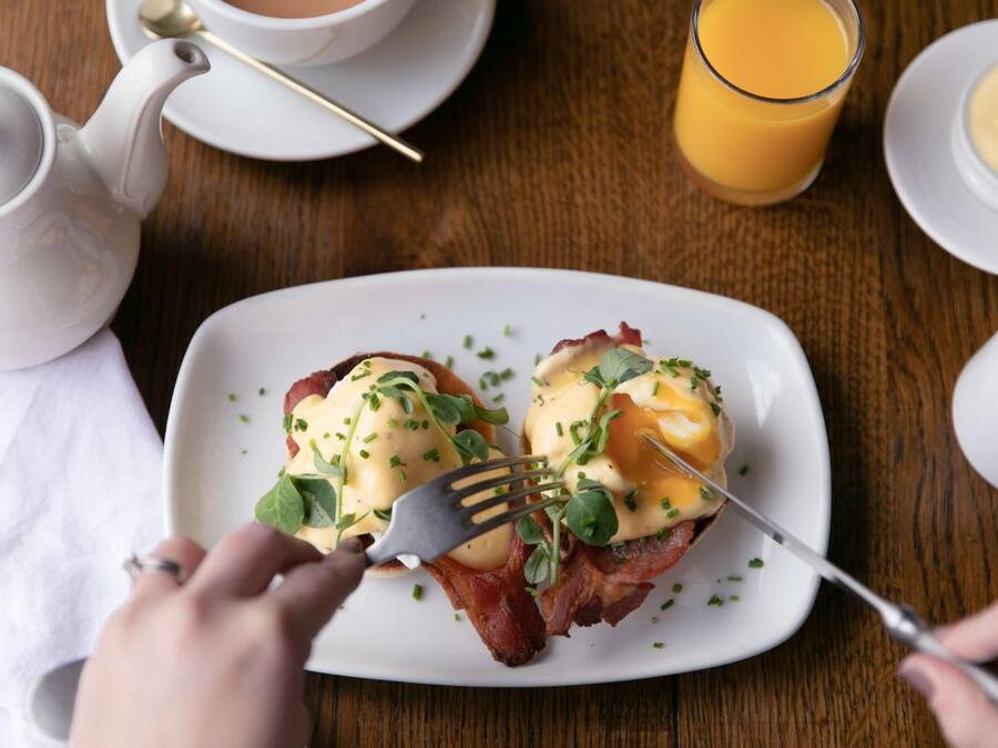 Un desayuno con un plato de huevos benedictinos, una tetera, un vaso de zumo de naranja y una taza de café.