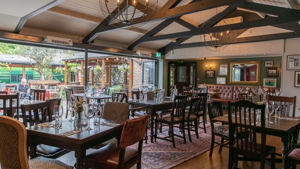 A restaurant with wooden tables and chairs, a chandelier, and a view of the outside. - The Rose & Crown Wimbledon (London)