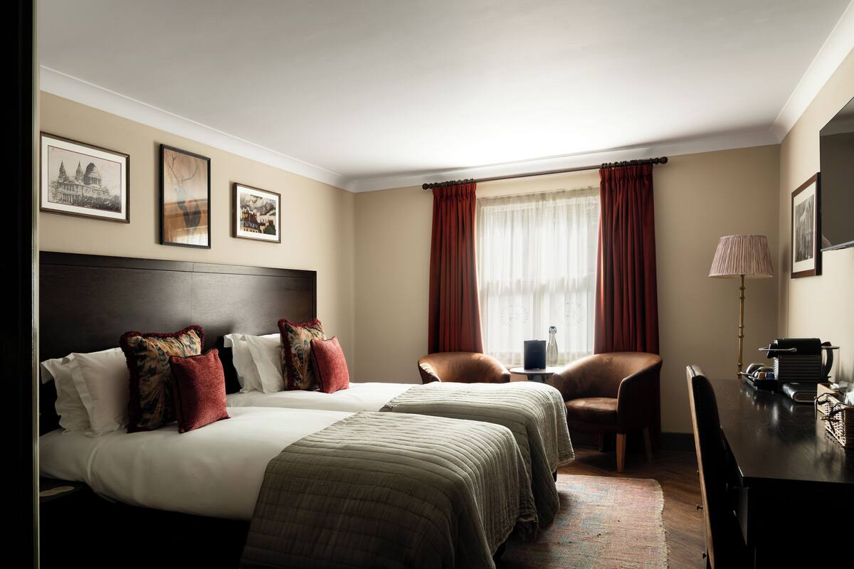 A hotel room with two beds, a desk, a chair, a lamp, and framed pictures on the wall.