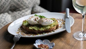 A plate with a poached egg on avocado toast, a fork, a knife, and a glass of white wine on a wooden table. - The Rose & Crown Wimbledon (London)