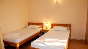Dos camas individuales con estructura de madera y sábanas blancas, una mesita de noche con lámpara entre ellas, y un aplique en la pared.