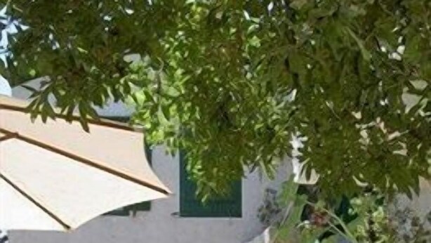 Un área de estar al aire libre con mesas y sillas, un paraguas grande y un edificio con fachada blanca y puerta arqueada.