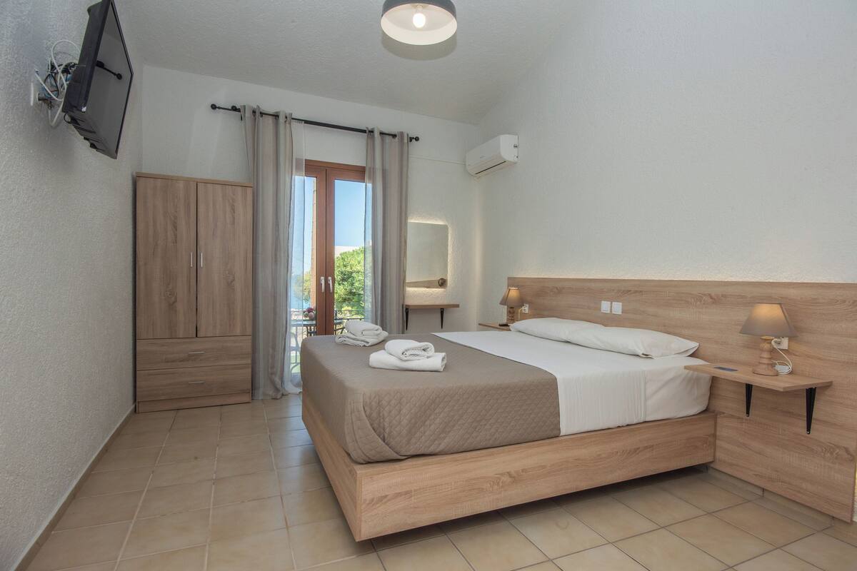 Ein Schlafzimmer mit Bett, Nachttisch, an der Wand montiertem Fernseher und einem Fenster mit Vorhängen.