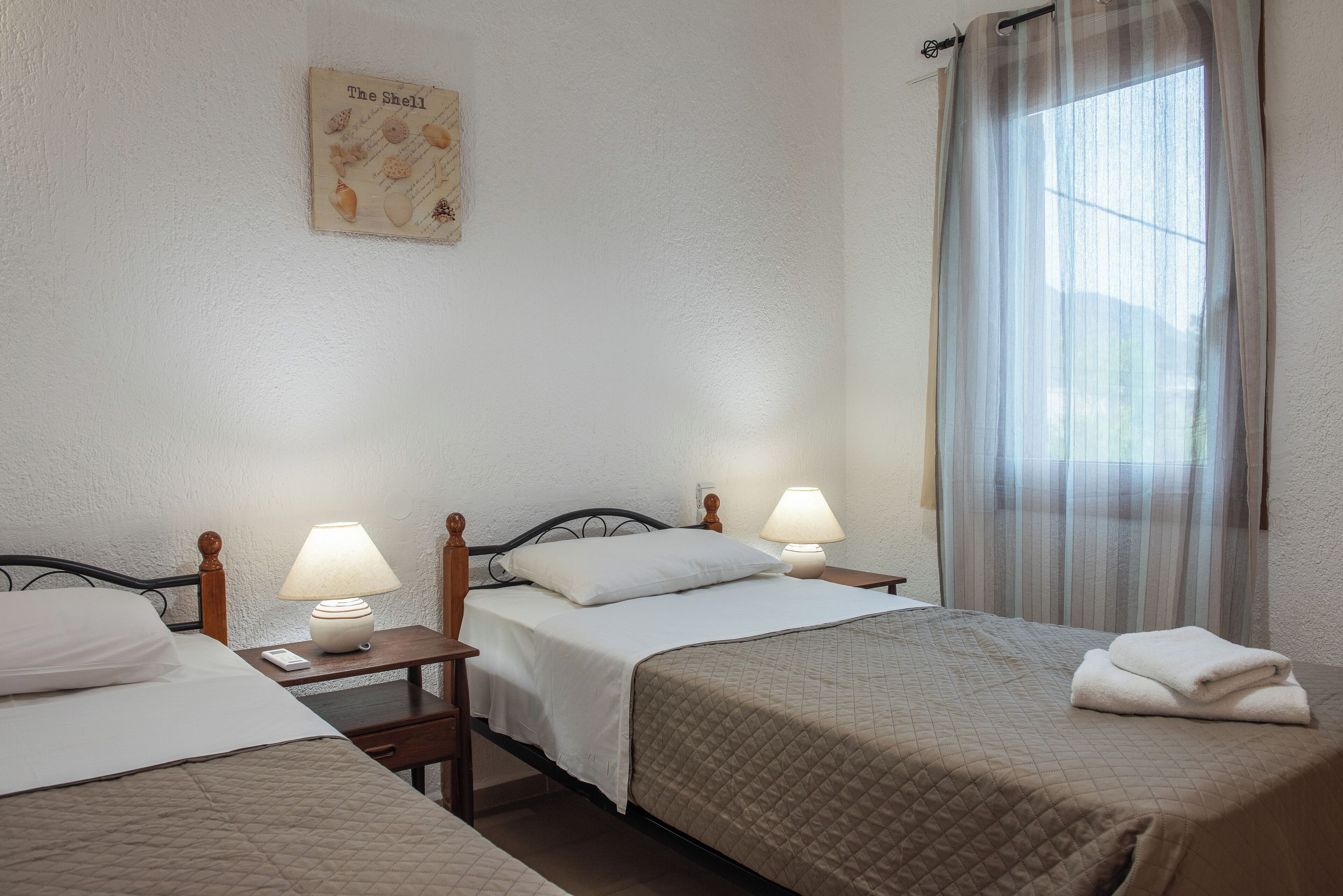 Een hotelkamer met twee bedden, elk met een nachtkastje en lamp, een raam met gordijnen, en een aan de muur hangend schilderij van schelpen.